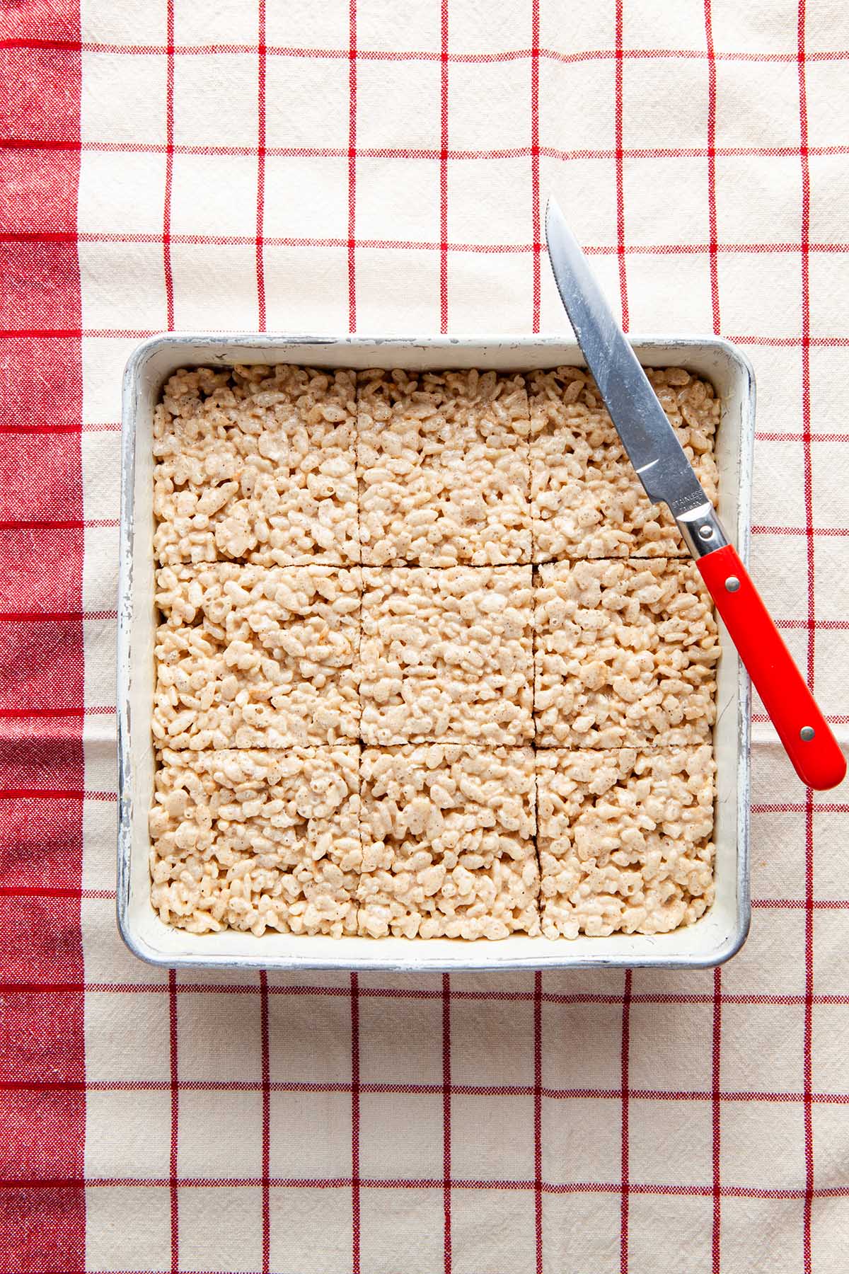 A pan of cut Rice Krispie squares with a red-orange-handled knife on a res checked tea towel.