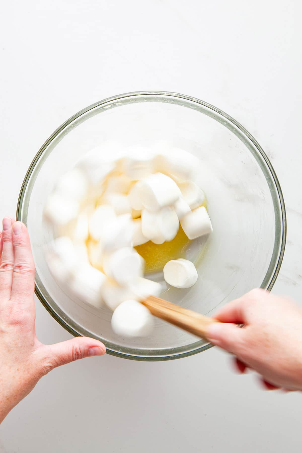 A hand stirring marshmallows and melted butter in a glass bowl with a rubber spatula.