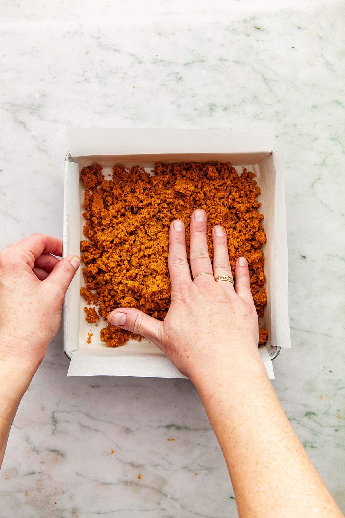 A hand beginning to press graham crumbs into a square pan.