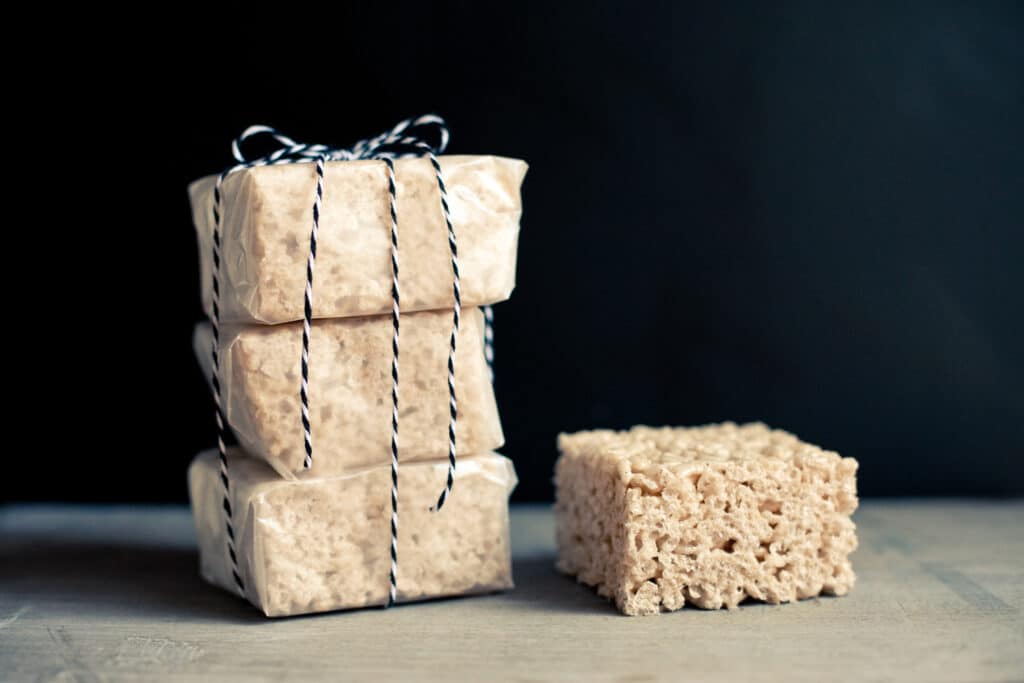 One chai-spiced rice krispie square on a board next to three wax paper-wrapped squares tied with twine.