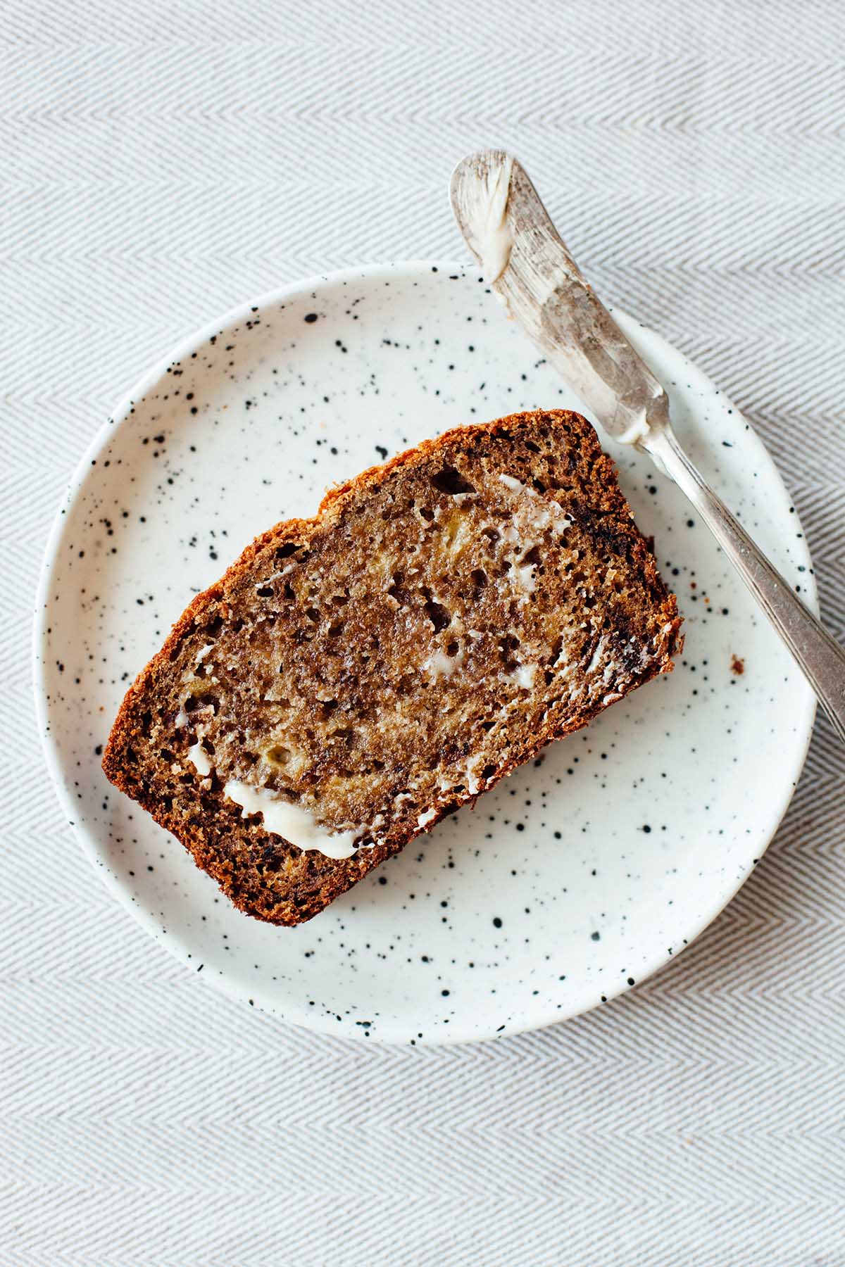 A slice of buttered banana bread on a small white speckled plate.