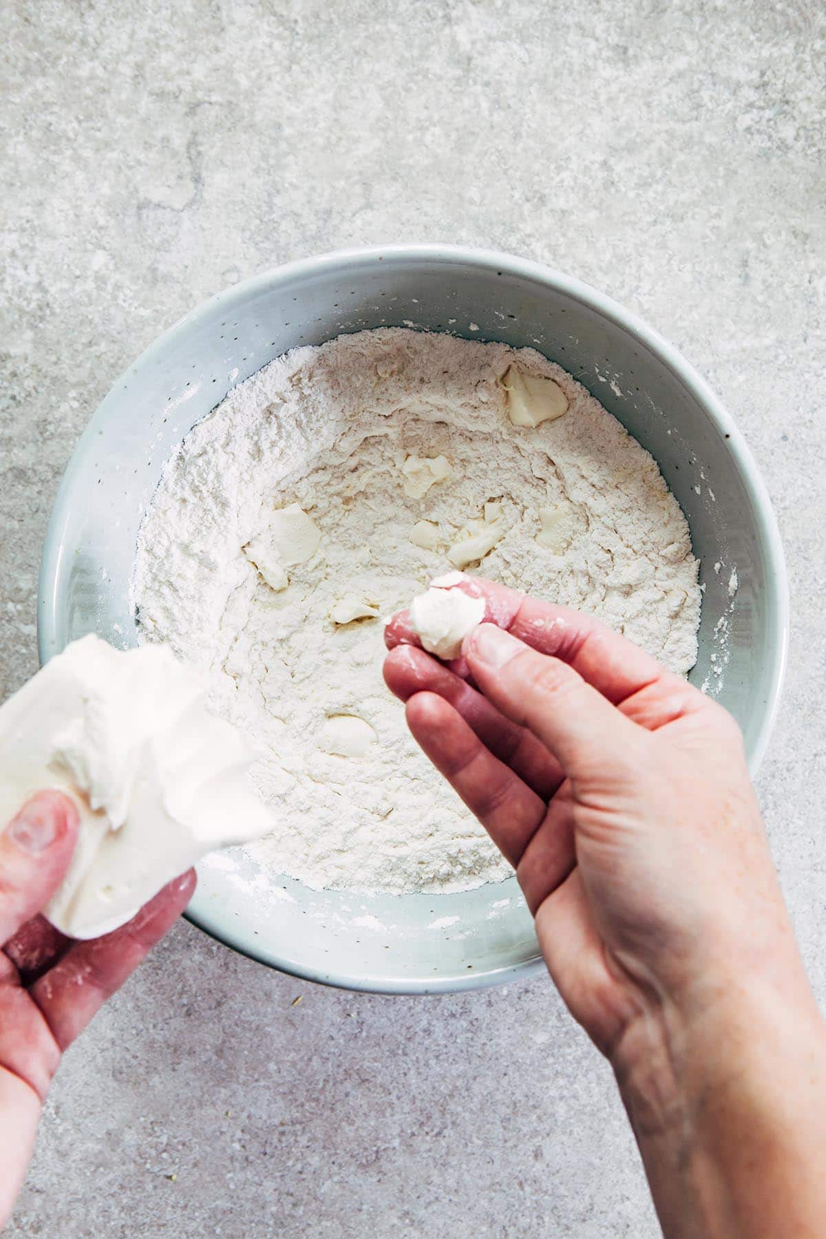 A hand breaking cream cheese into small pieces and placing them in a bowl of flour.