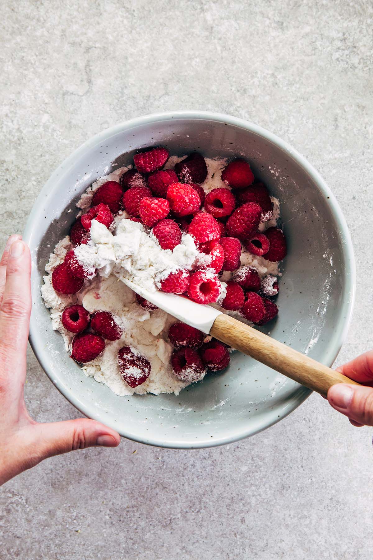A hand mixing dry muffin ingredients with raspberries using a rubber spatula before the liquid has been added.
