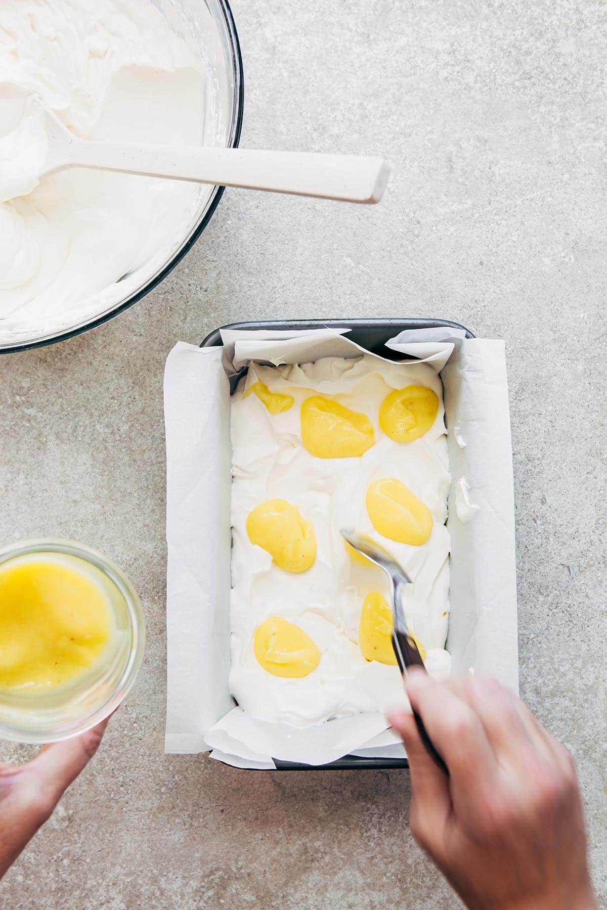 A hand dolloping lemon curd all over a loaf tin filled with whipped cream.