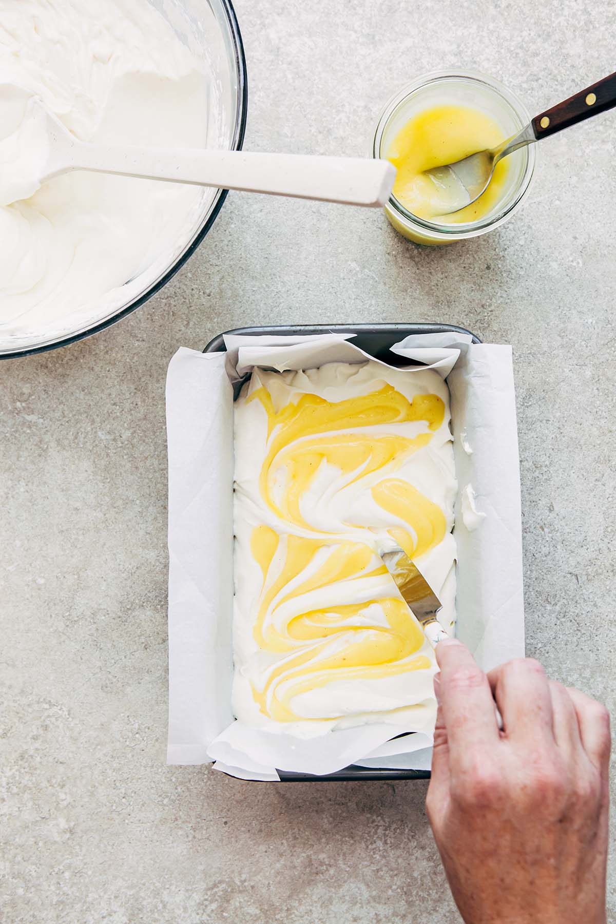 A hand swirling lemon curd through a tin of no-churn ice cream.