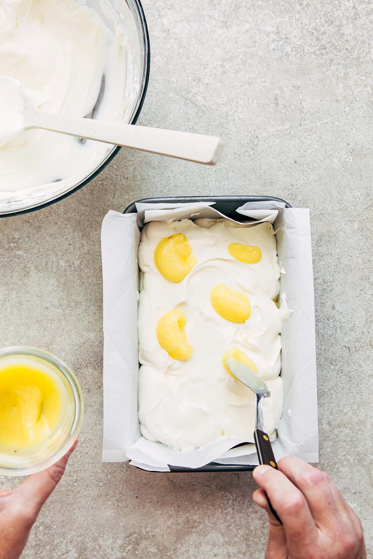 A hand spooning lemon curd into a pan of whipped cream.
