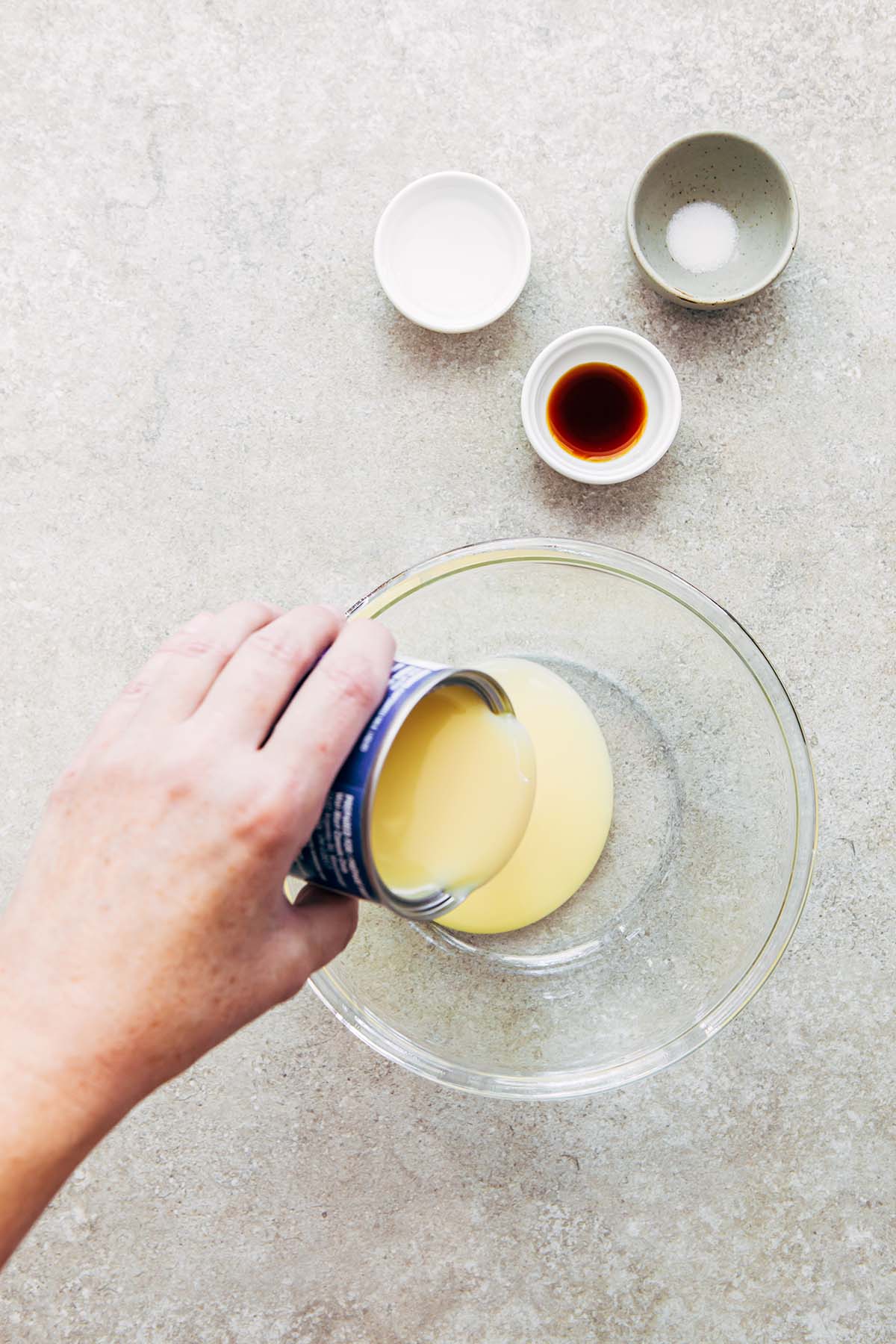 A hand pouring a can of condensed milk into a glass bowl.