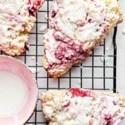 A wire cooling rack topped with glazed raspberry scones and a small pink bowl of lemon glaze.