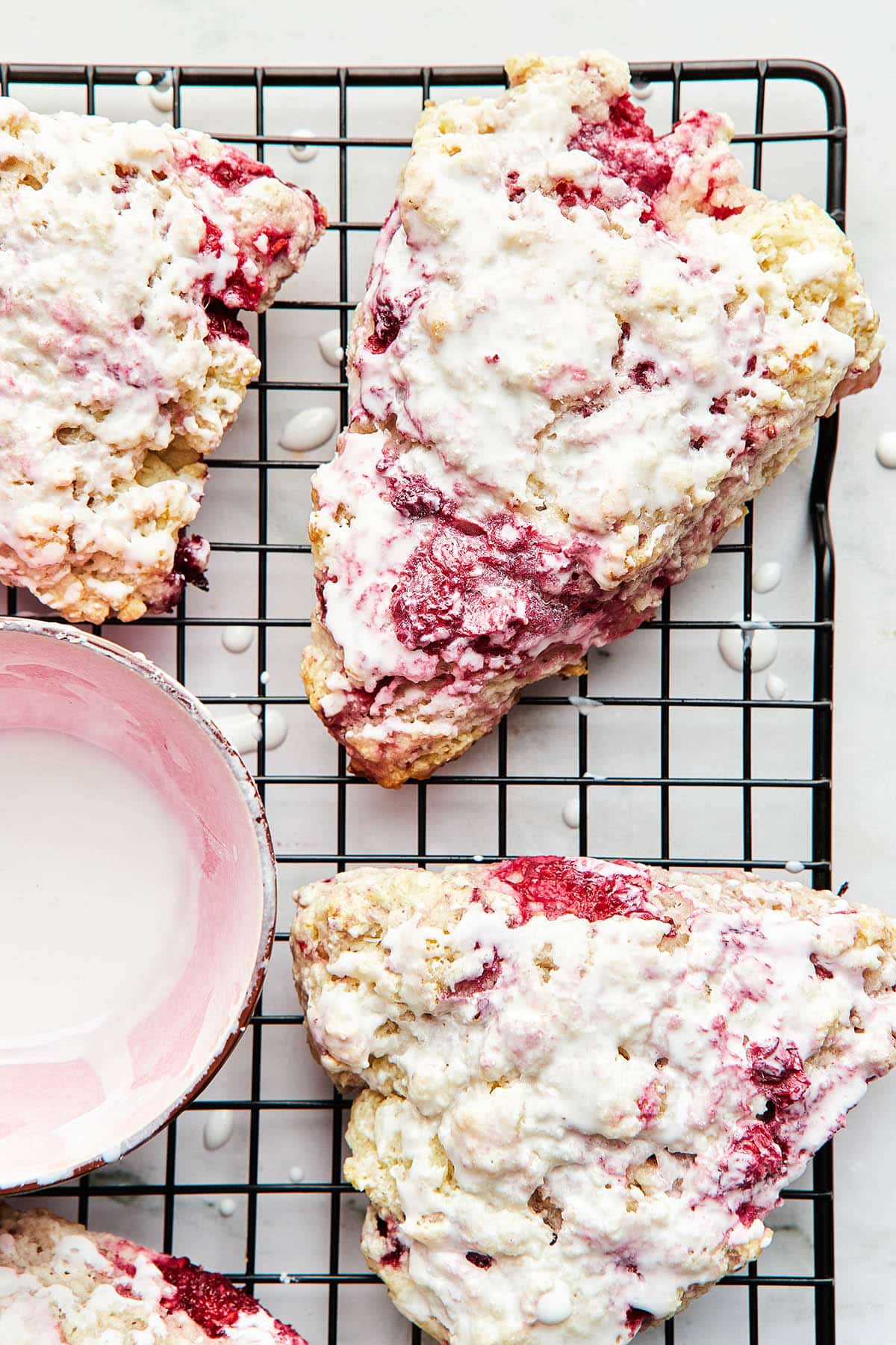 A wire cooling rack topped with glazed raspberry scones and a small pink bowl of lemon glaze.