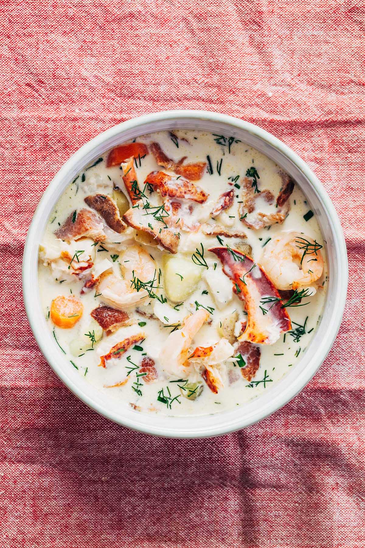 Overhead shot of a bowl of nova Scotia seafood chowder loaded with lobster, haddock, shrimp, potatoes, and bacon, with chopped dill and chives on a red fabric background.