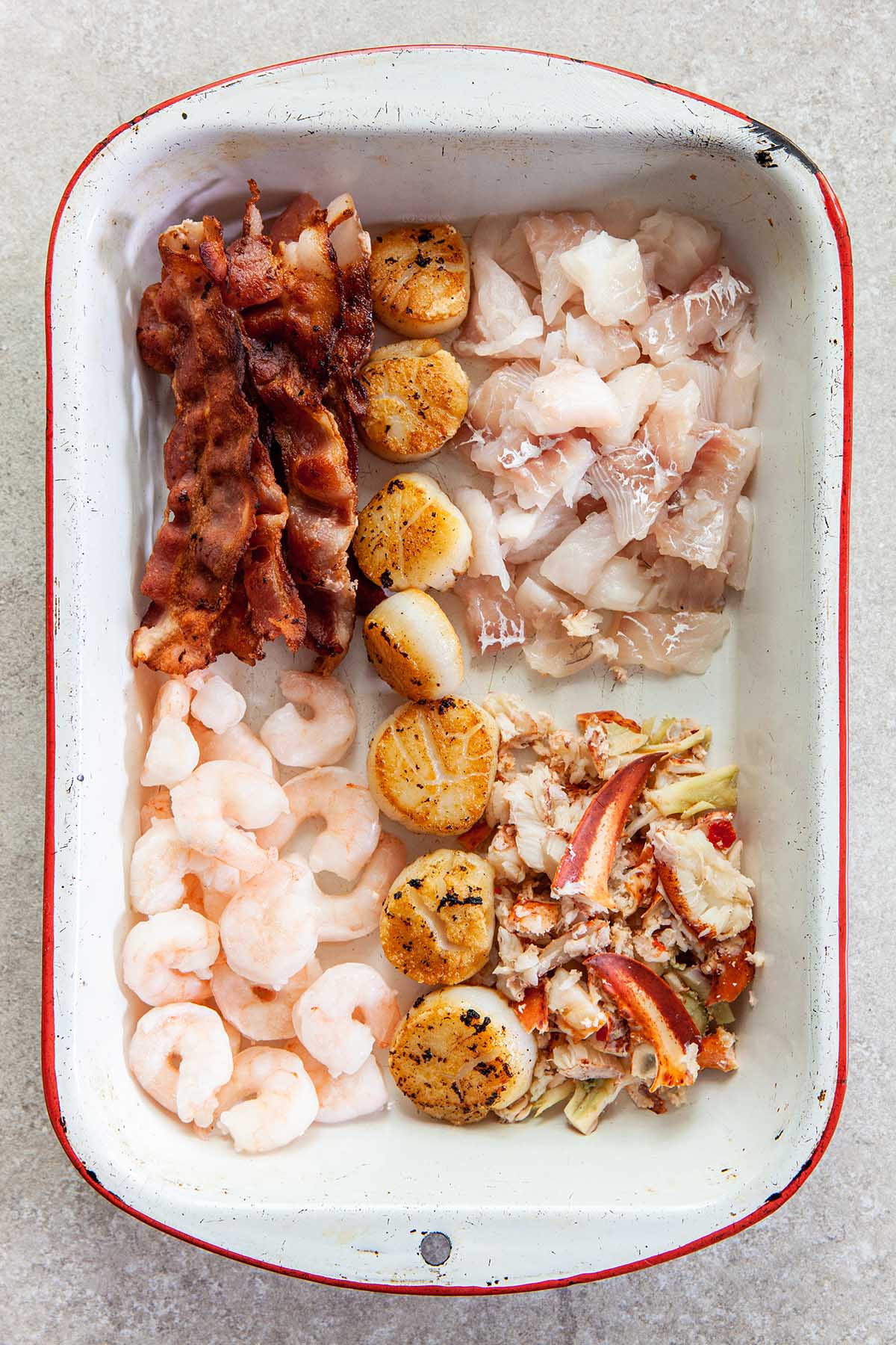 A large white enamel baking dish with red trim filled with slices of cooked bacon, pan-seared scallops, fresh haddock pieces, cooked lobster meat, and small cooked shrimp.