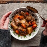 A bowl of Dutch oven beef stew on linen atop a rustic wooden table, with a woman’s hands holding the bowl and a spoon.