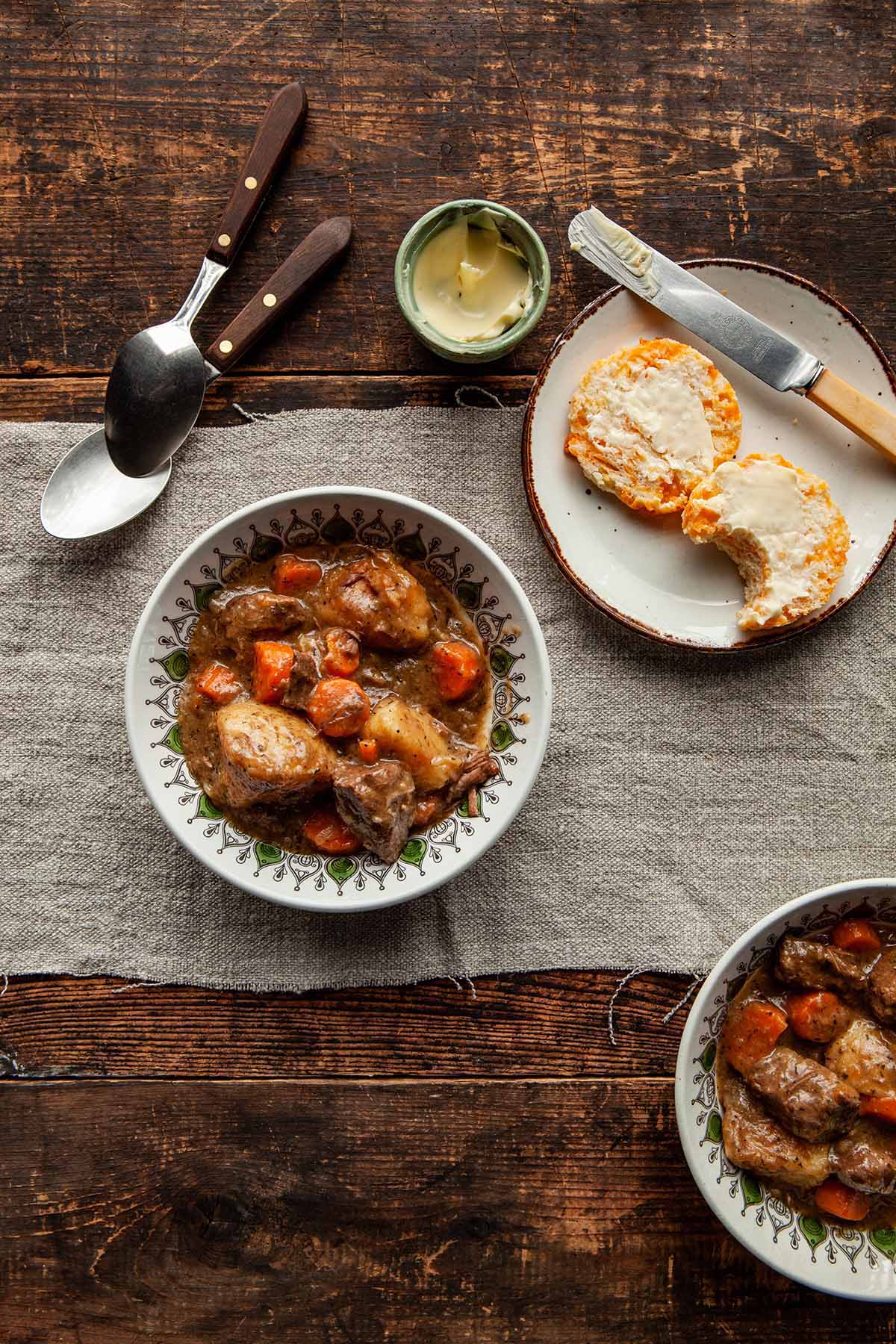 Two bowls of Dutch oven beef stew on a wood table. A small dish of butter and a cheese biscuit, split in half and buttered, with one bite taken, on a plate nearby.