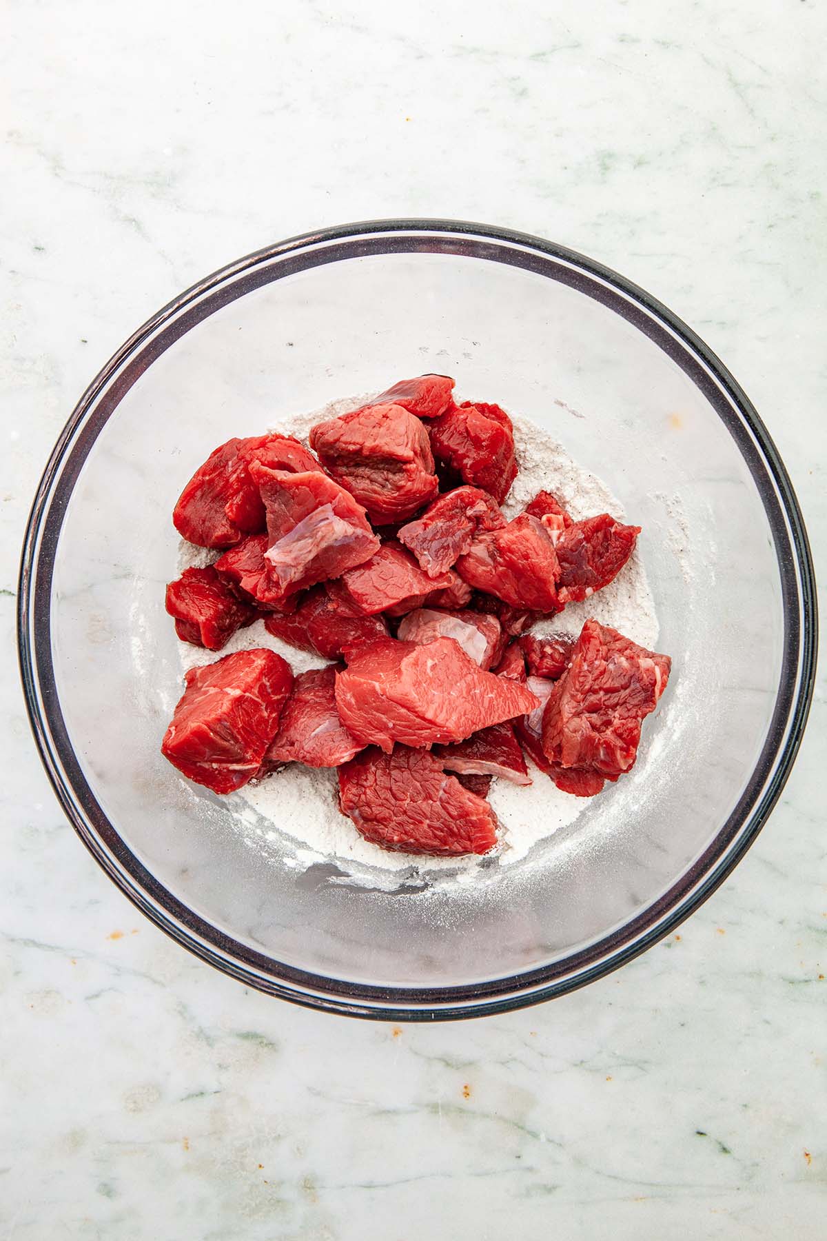 A glass bowl of flour, salt, and pepper with beef stewing cubes on top.