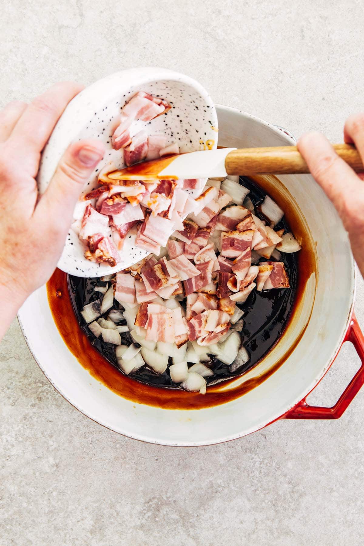 Hands scraping chopped bacon from a bowl into a pot of onion and molasses mixture.