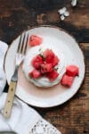 Single pavlova shell topped with watermelon on a pottery plate on a rustic table.