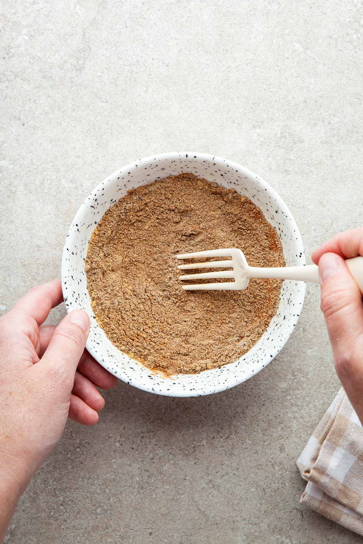 A hand using a fork to mix spices in a bowl.