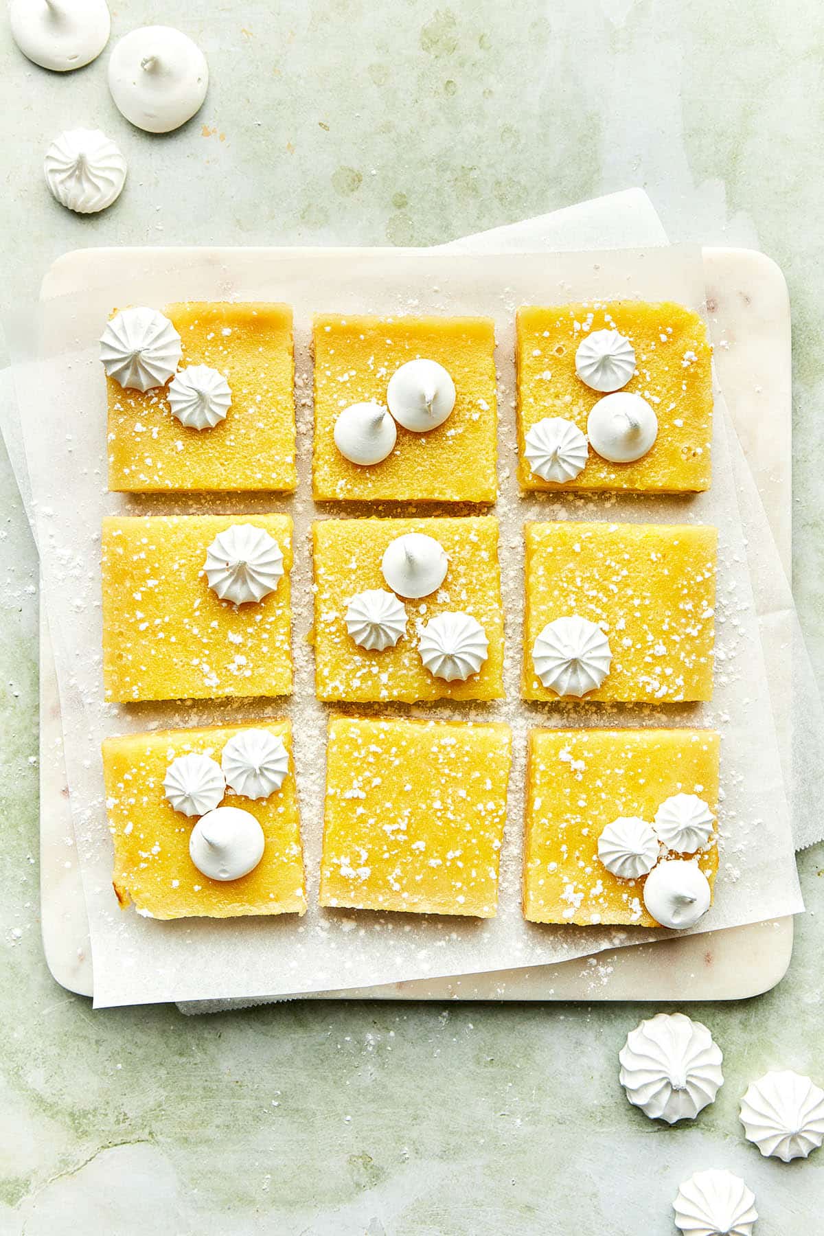 Nine sliced lemon squares on a marble board sprinkled with powdered sugar and topped with mini piped meringues.