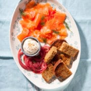 A platter of salmn gravlax, pickled red onion, toasted brown bread, and aioli with sesame seeds.