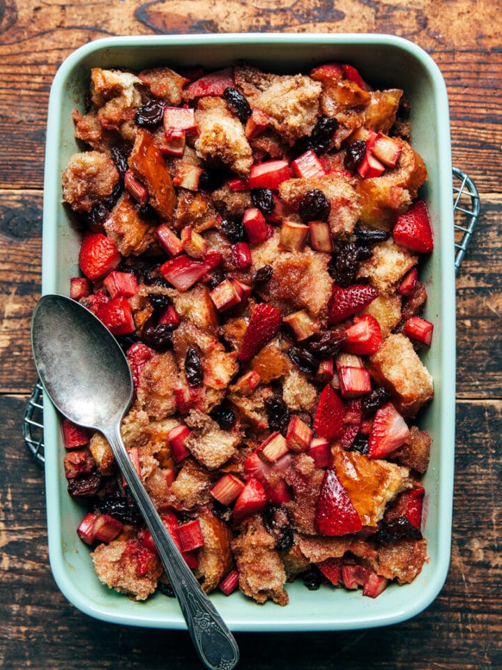 A green baking dish filled with strawberry rhubarb bread pudding with a vintage serving spoon on a rustic farmhouse table.