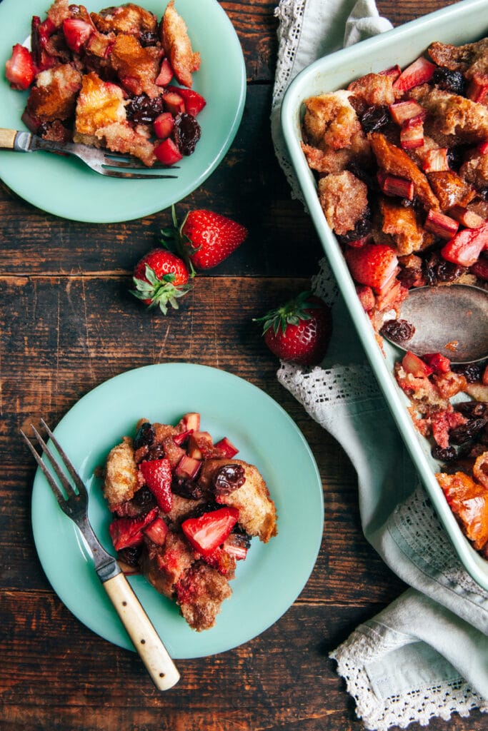 Two plates of strawberry rhubarb bread pudding next to the baking dish on a rustic harvest table.