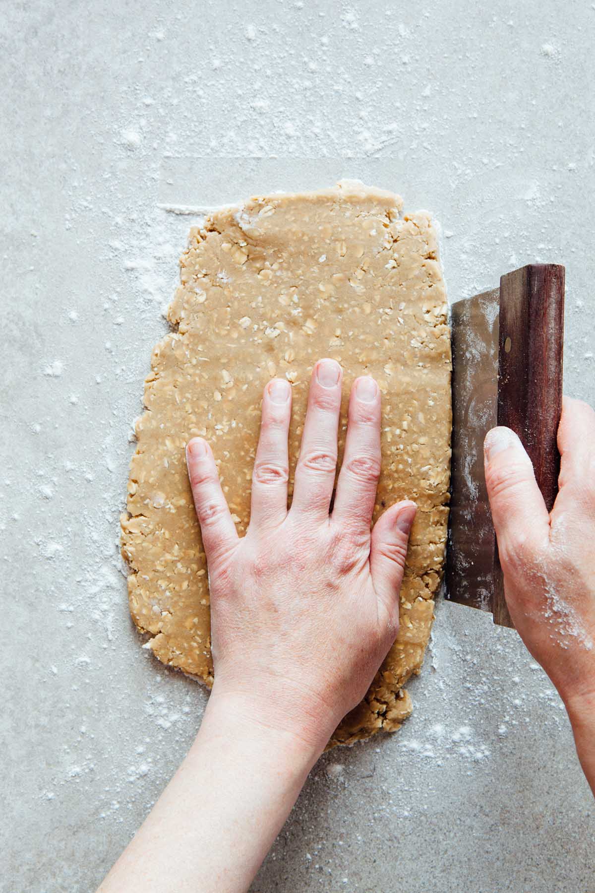 A hand squaring off oatcake dough along the sides with a bench scraper.
