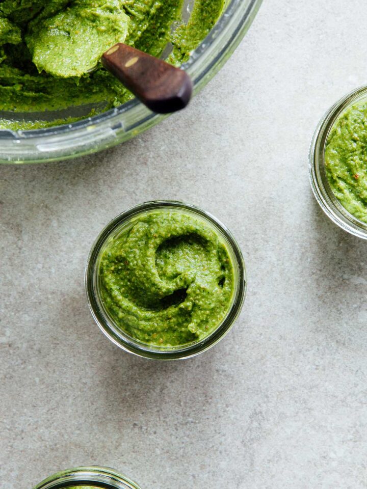 Small jars of spinach arugula pesto next to the bowl of a food processor with more pesto and a spoon inside of it.