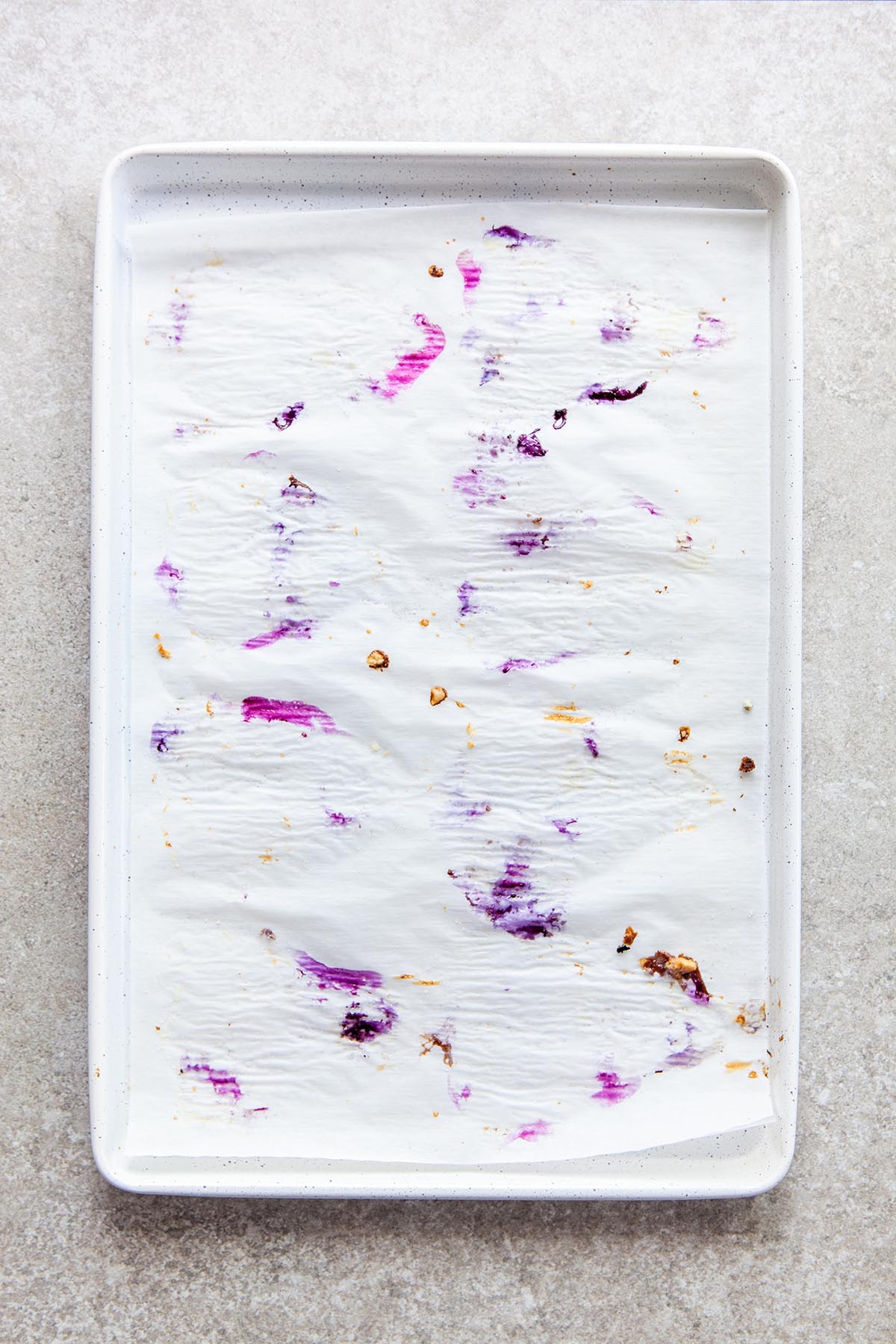 A white baking sheet lined with cooked blueberry-spattered parchment paper on a stone surface.