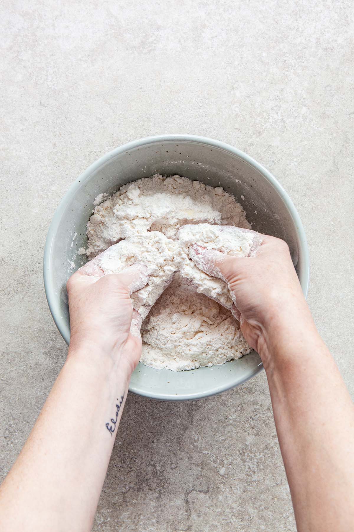Two hands rubbing butter into flour inside a light blue bowl.