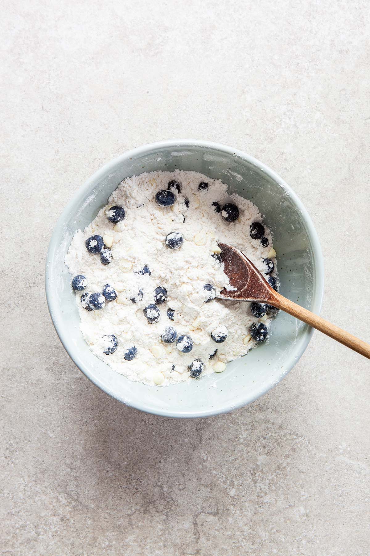 A bowl with flour, blueberries, and white chocolate chips just stirred with a wooden spoon that's inside the bowl.