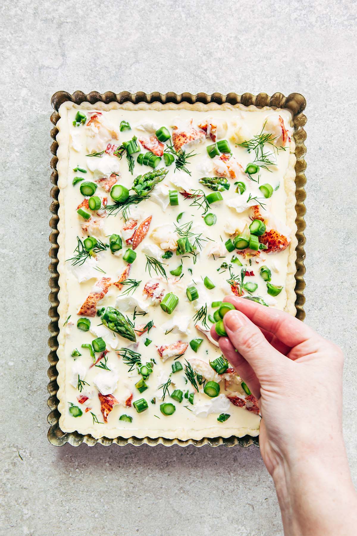A woman's hand sprinkling sliced asparagus over an unbaked lobster quiche in a rectangular metal tart tin on a stone surface.