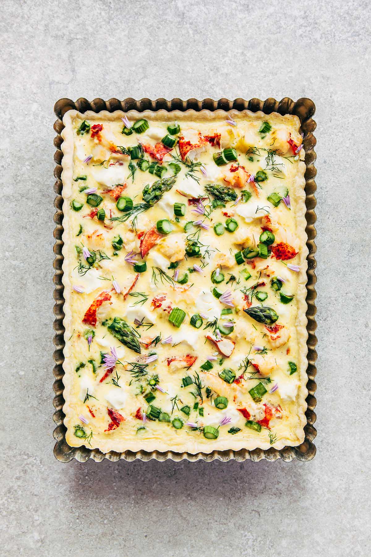 Overhead close up of a lobster quiche with cream cheese and asparagus in a rectangular metal tart tin on a stone surface.