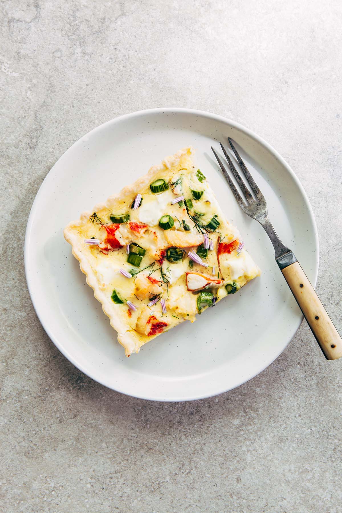 Overhead shot of one square slice of lobster quiche on a white plate with a fork, all on a stone surface.