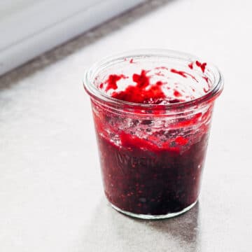 A small glass jar of raspberry rhubarb jam on a stone surface next to a white windowsill.