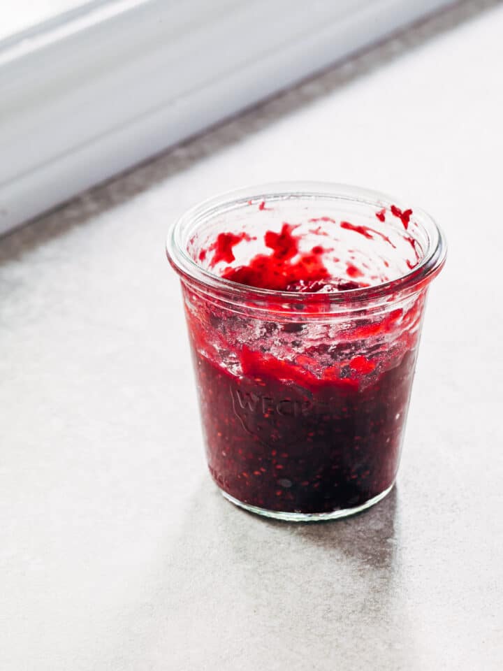 A small glass jar of raspberry rhubarb jam on a stone surface next to a white windowsill.