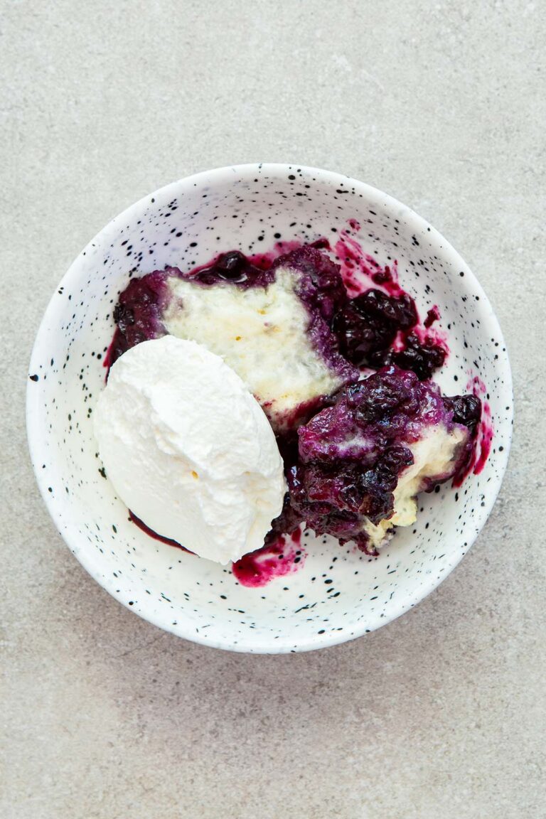 A serving of Nova Scotia blueberry grunt topped with whipped cream in a white speckled bowl on a stone surface.