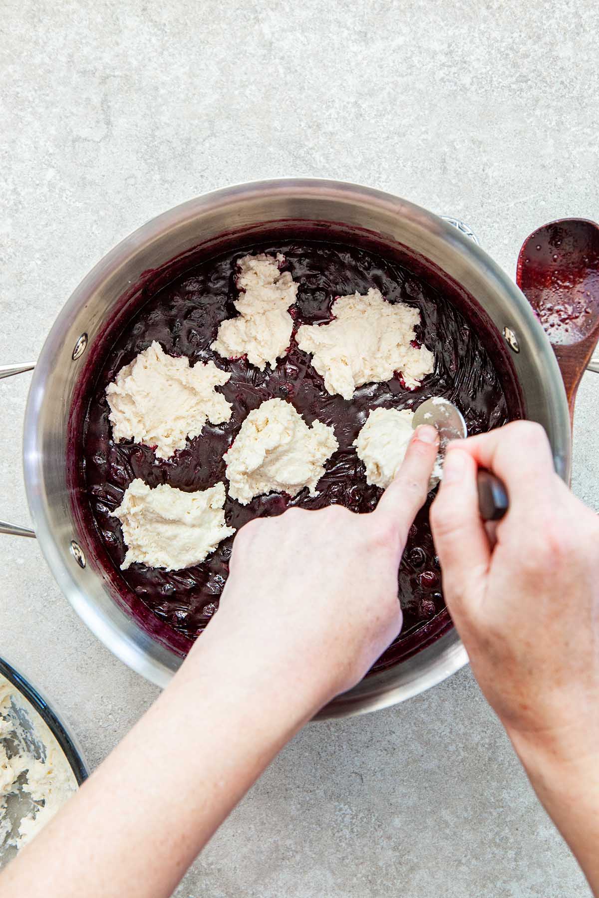 Hands scraping biscuit dough from a spoon into a pot of cooked blueberries.