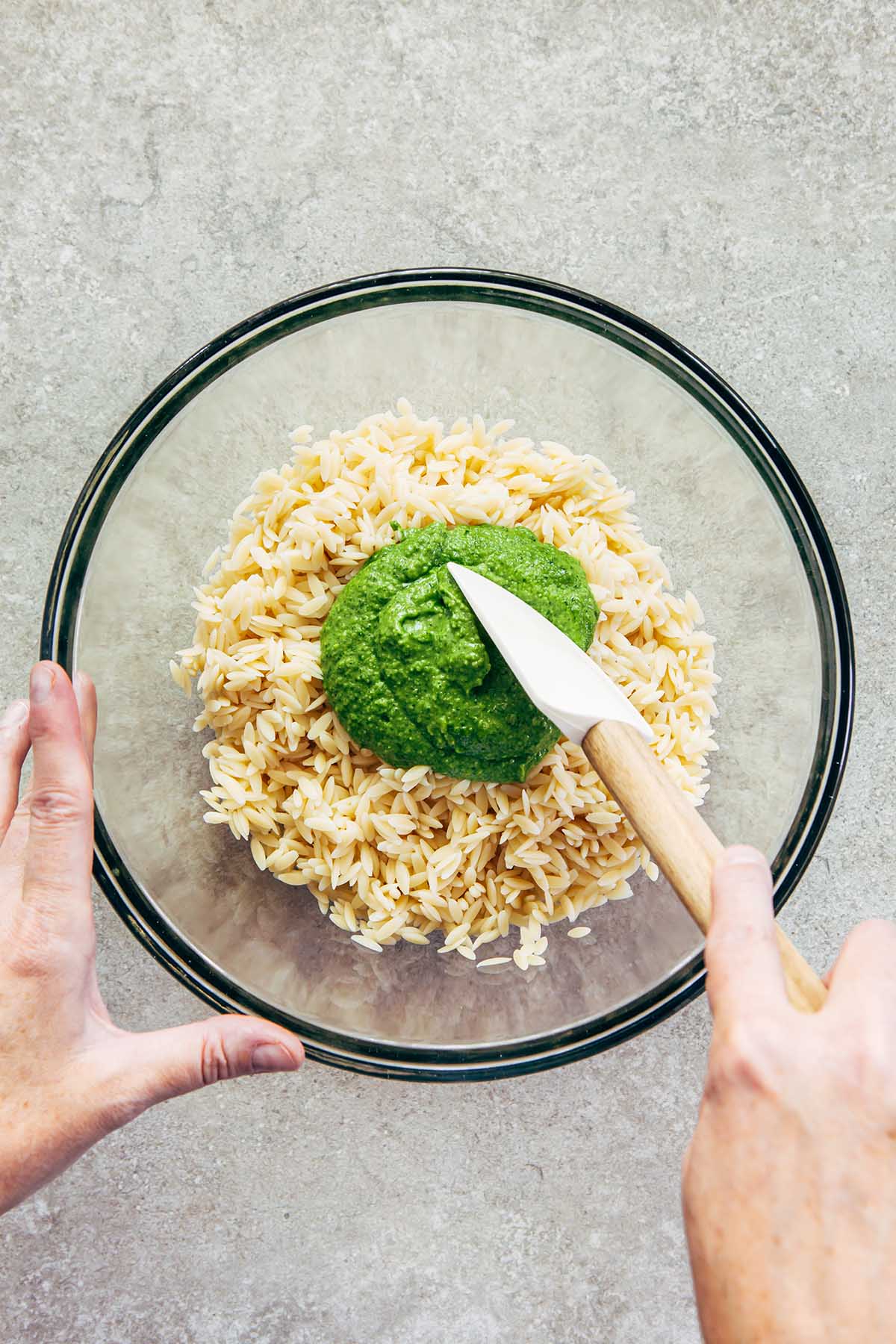 Hands stirring green pesto into a bowl of cooked orzo.