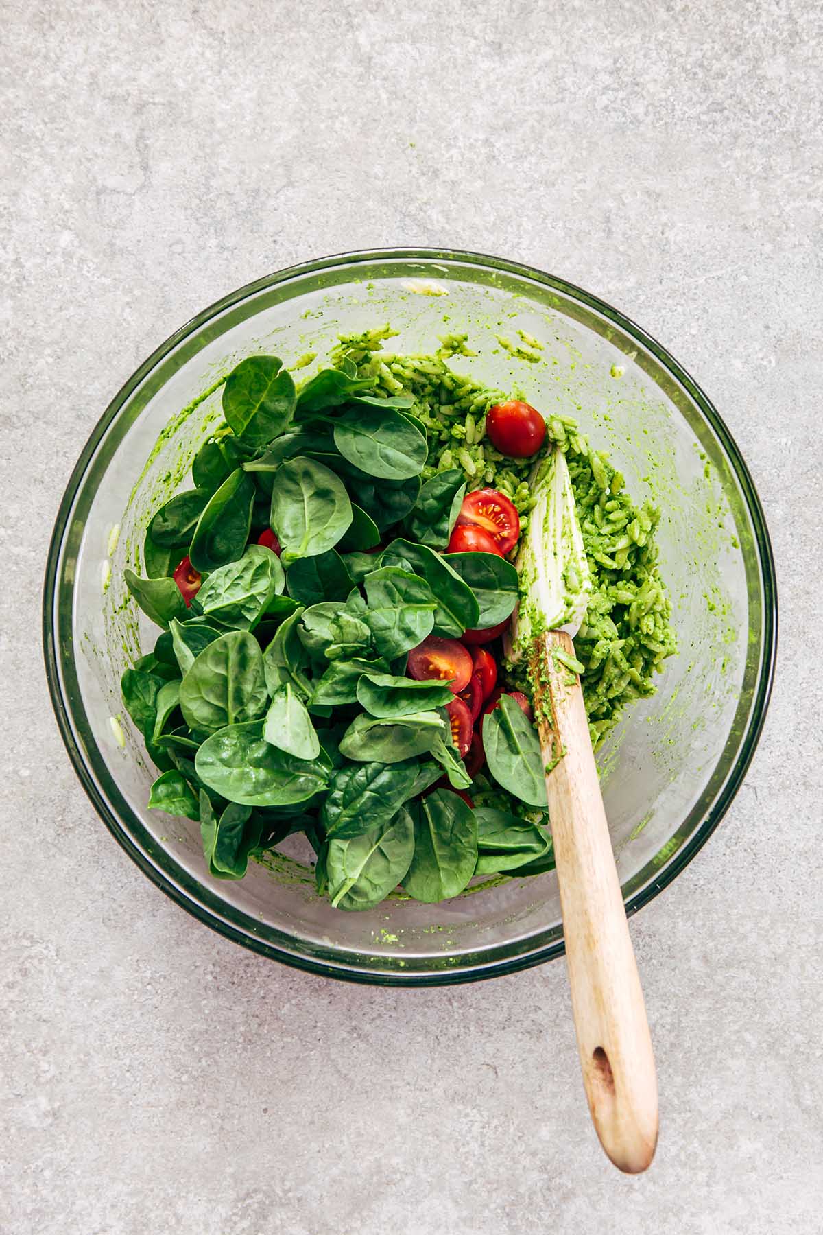 A glass bowl of pesto orzo, halved cherry tomatoes, baby spinach leaves, and torn basil.