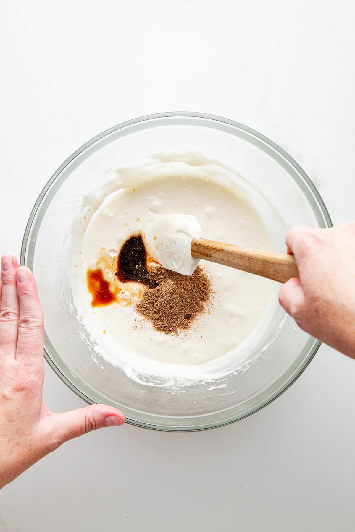 A hand about to mix melted marshmallows, spices, and vanilla in a glass bowl with a rubber spatula.