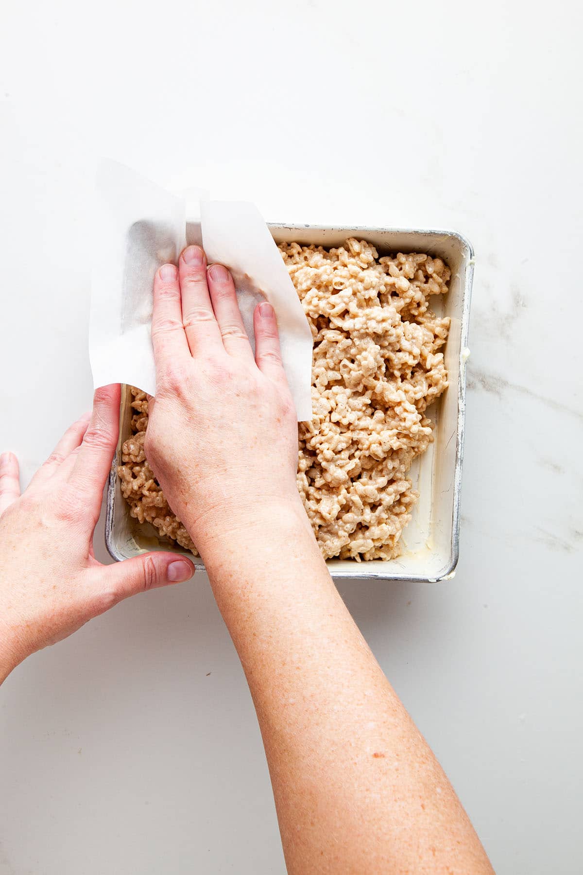 A hand smoothing the top of a pan of marshmallow treats with a small piece of parchment paper.