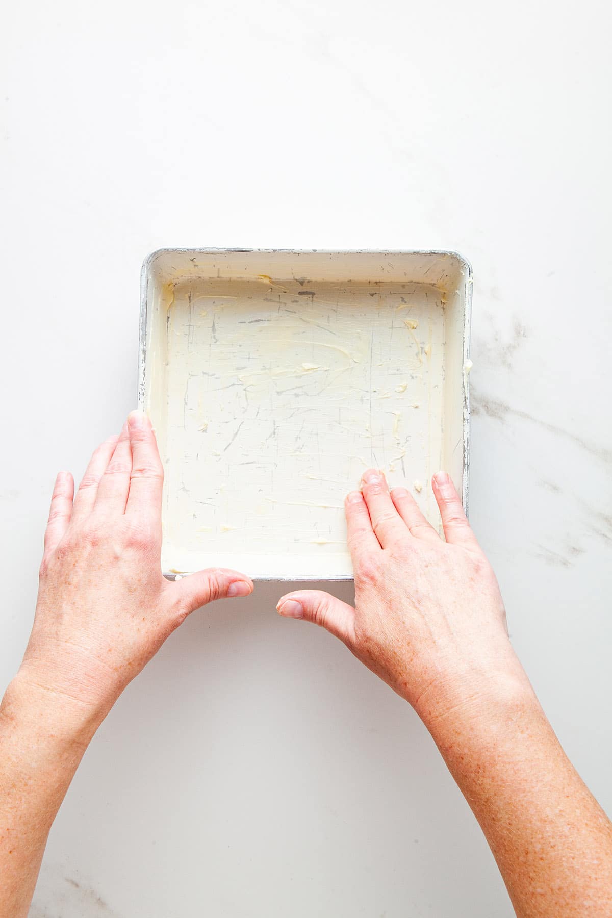 A hand smearing butter on the inside of a square baking pan.