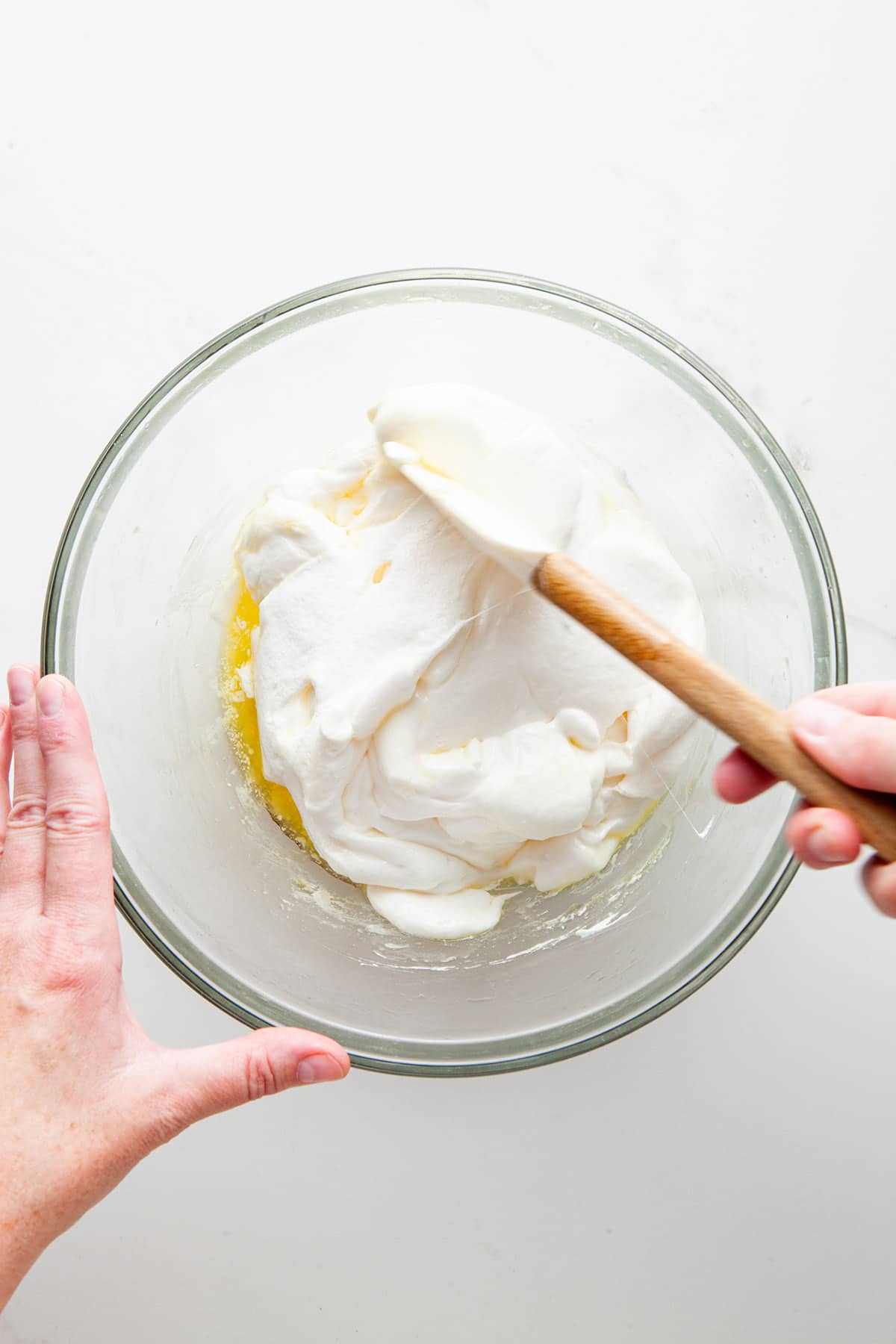 A hand stirring melted marshmallow and butter in a glass bowl with a rubber spatula.
