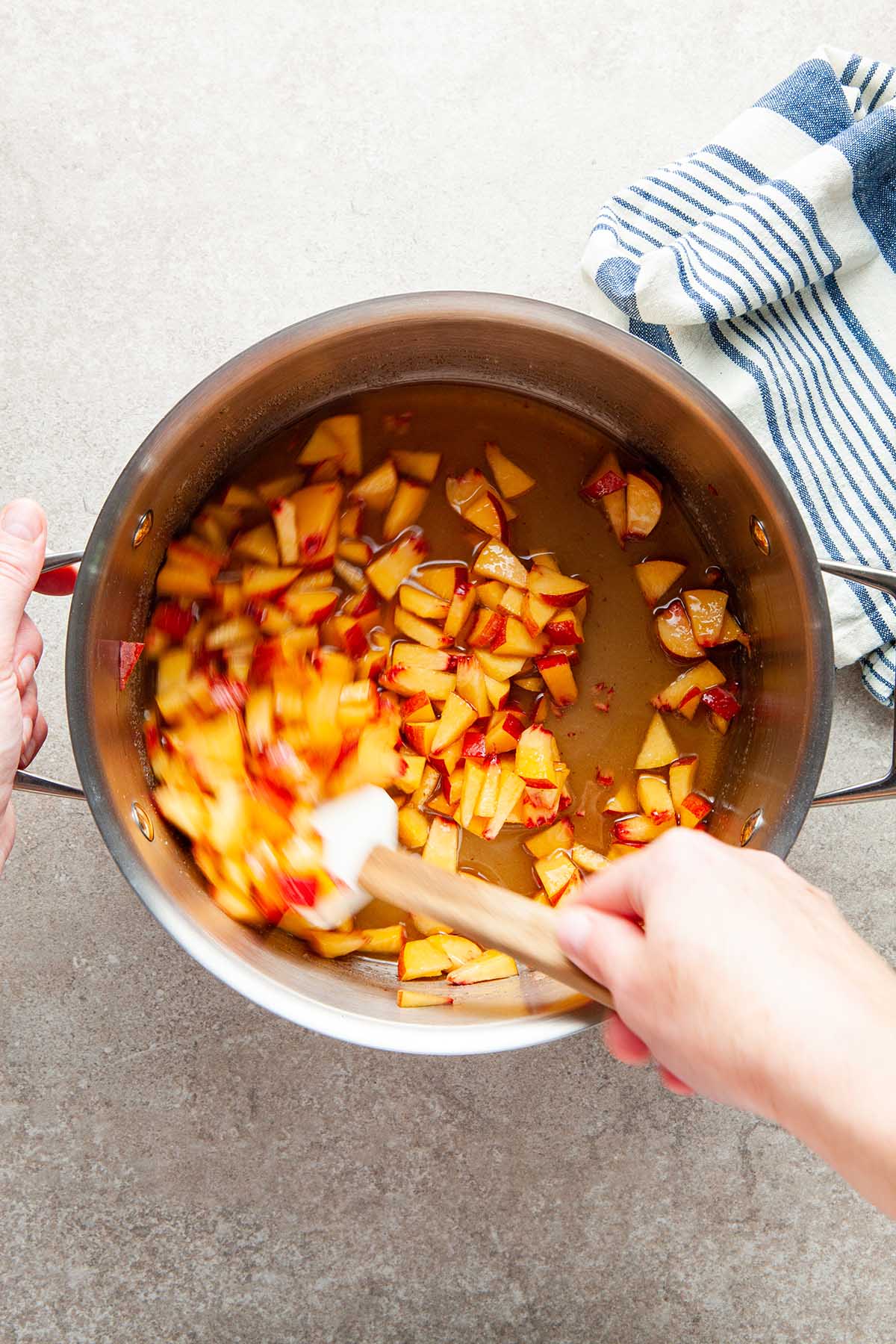 A hand stirring peaches, maple syrup, and sugar together in a pot with a rubber spatula.