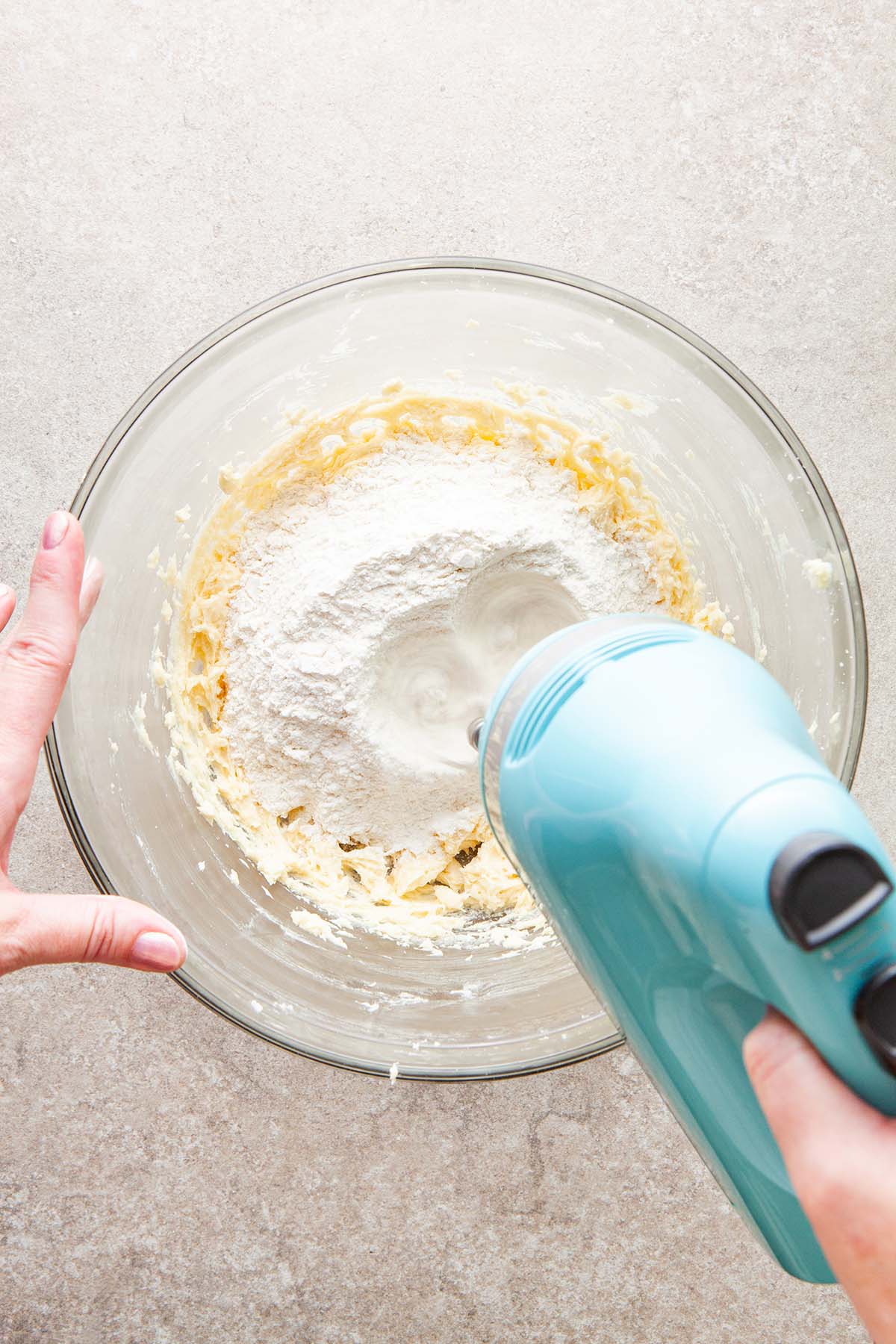 A hand holding a hand mixer to mix flour into butter in a glass bowl.