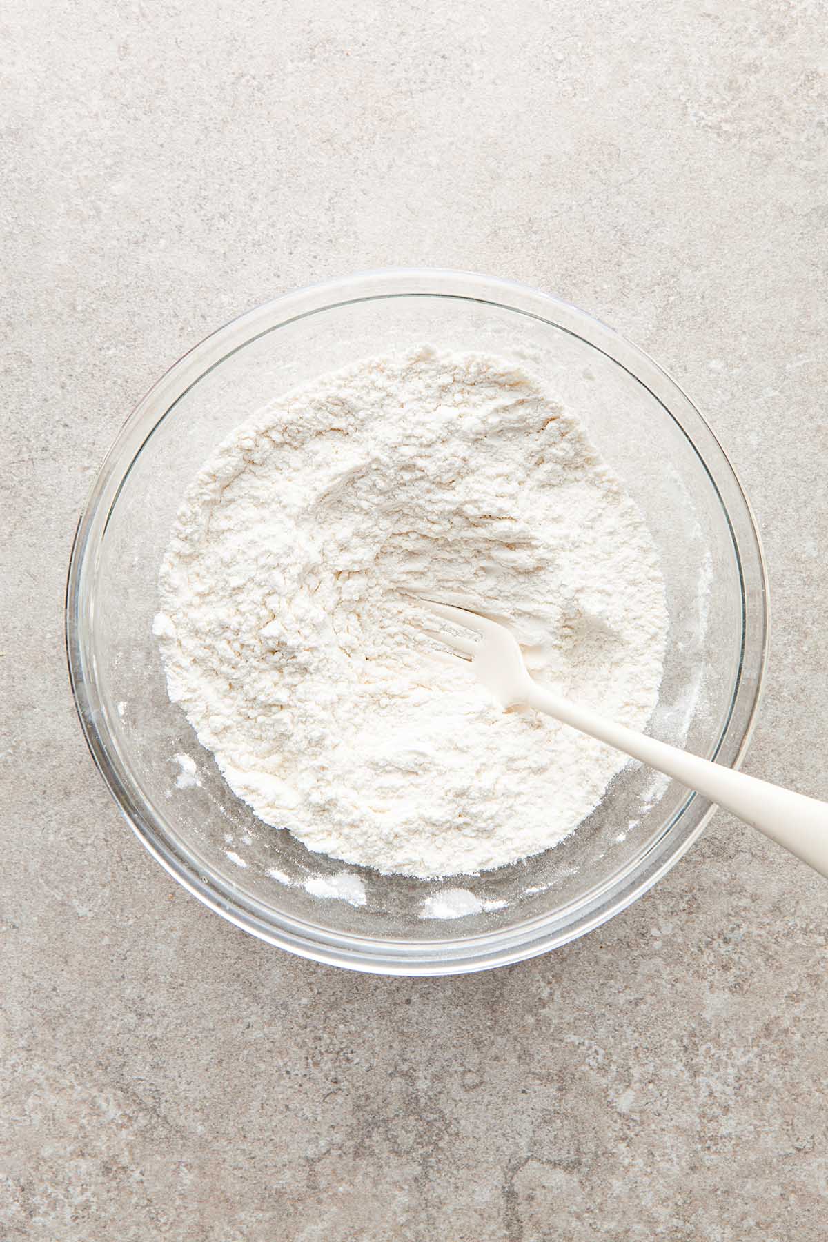 A bowl of flour, baking poweder, baking soda, and salt mixed with a fork.
