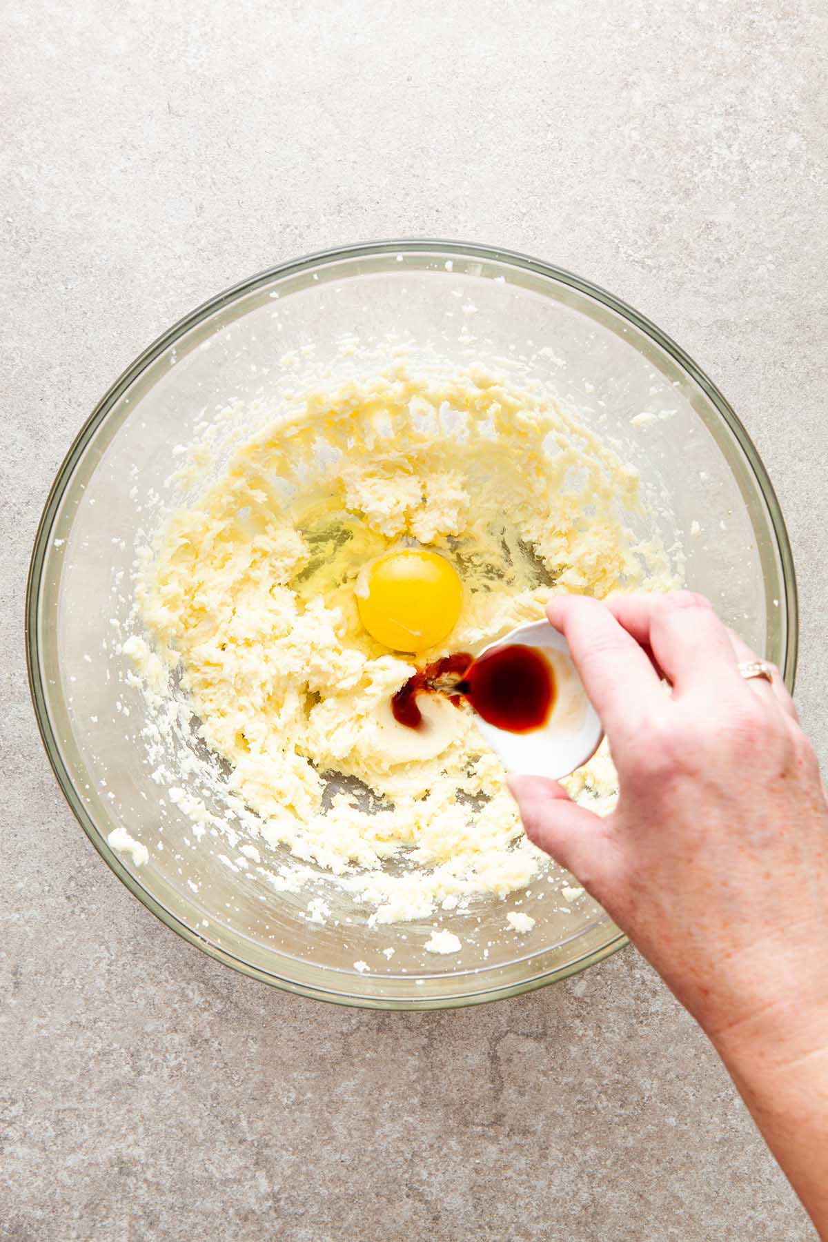 A hand pouring vanilla extract from a small bowl into a large glass bowl of creamed butter and egg.