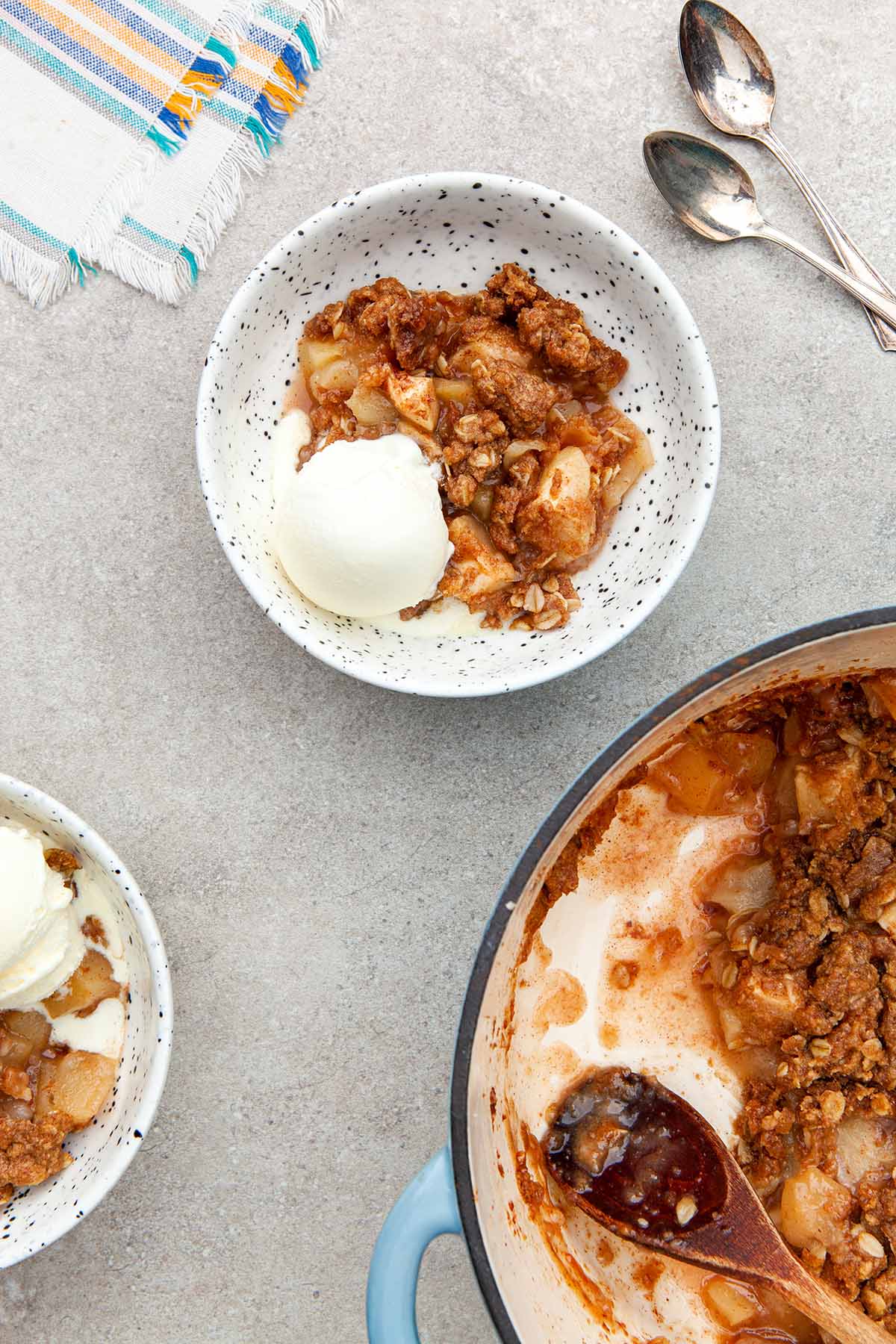 Two servings of Dutch oven apple crisp with ice cream in bowl on a stone surface next to the pot.