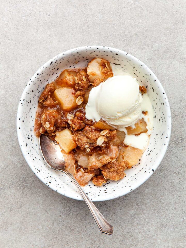 A bowl of apple crisp topped with ice cream in a speckled bowl.