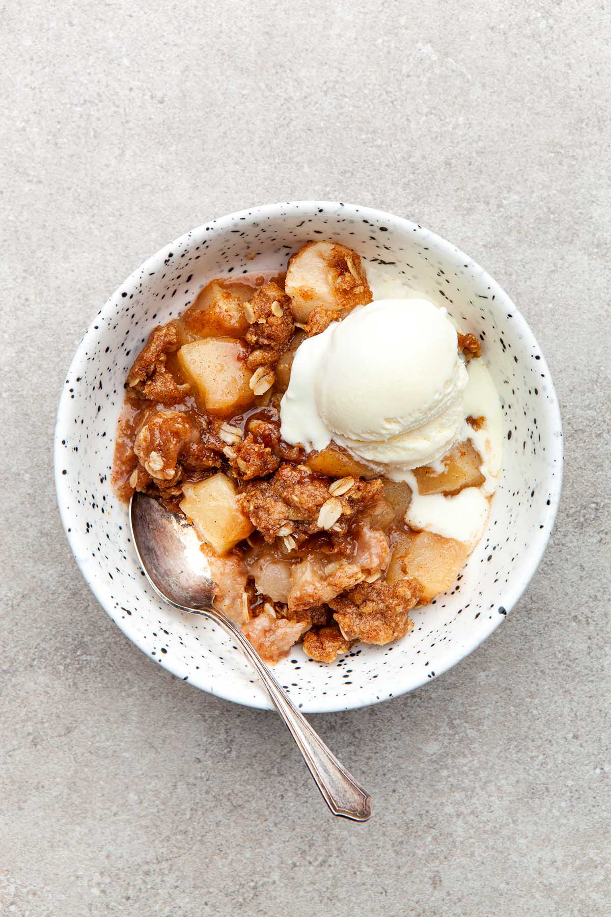 A bowl of apple crisp topped with ice cream in a speckled bowl.